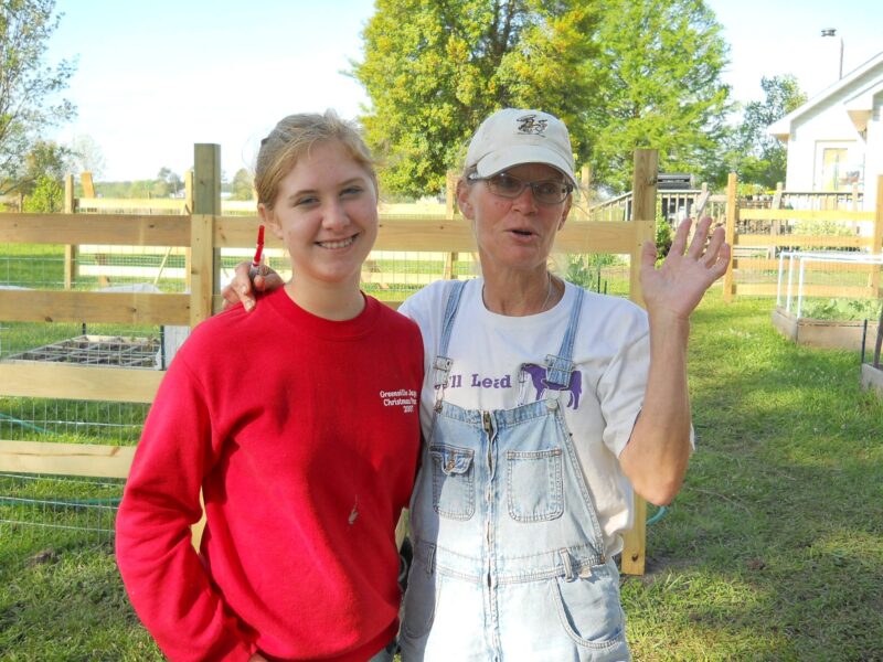 Two women smiling together in a garden setting, with wooden fences and vegetable beds in the background. One woman wears a red sweatshirt and holds a pen, while the other is in overalls and a cap, gesturing with her hand. The scene captures a friendly and welcoming atmosphere in an outdoor farming environment.