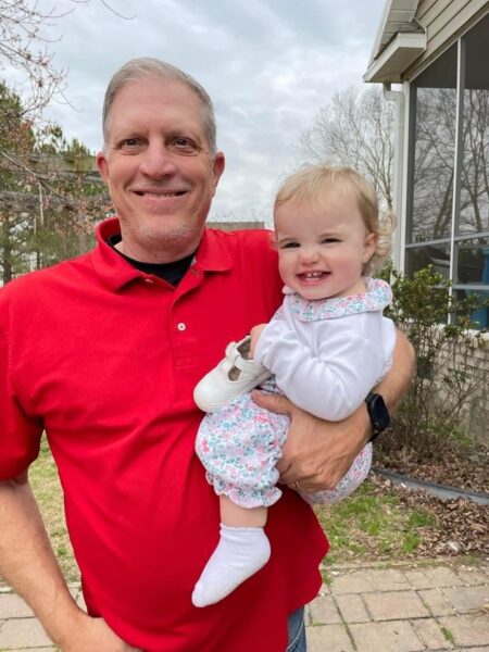 Smiling grandfather holding a happy baby girl outdoors, both wearing casual clothing. The grandfather is in a red shirt, and the baby is dressed in a floral outfit with white shoes. The background features a house and trees under a cloudy sky.