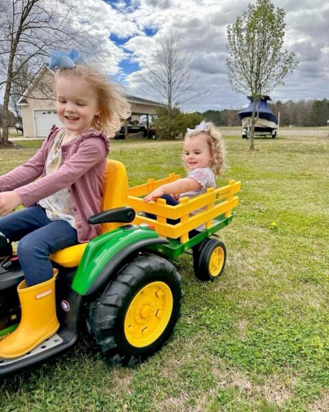 Two young girls enjoy a sunny day outdoors, riding on a green toy tractor with a yellow wagon in tow. The older girl, wearing a pink cardigan and yellow boots, steers the tractor while the younger girl, dressed in a light pink outfit, sits in the wagon smiling. The background features a house and trees under a partly cloudy sky, highlighting a cheerful family activity.