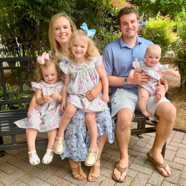 A happy family of five sits on a park bench surrounded by greenery. The mother, wearing a floral dress, holds one daughter, while another daughter, dressed in matching floral attire with a pink bow, sits beside her. The father, in a blue polo shirt, holds their baby, who also wears a floral outfit. The scene captures a joyful moment of family bonding in a natural setting.