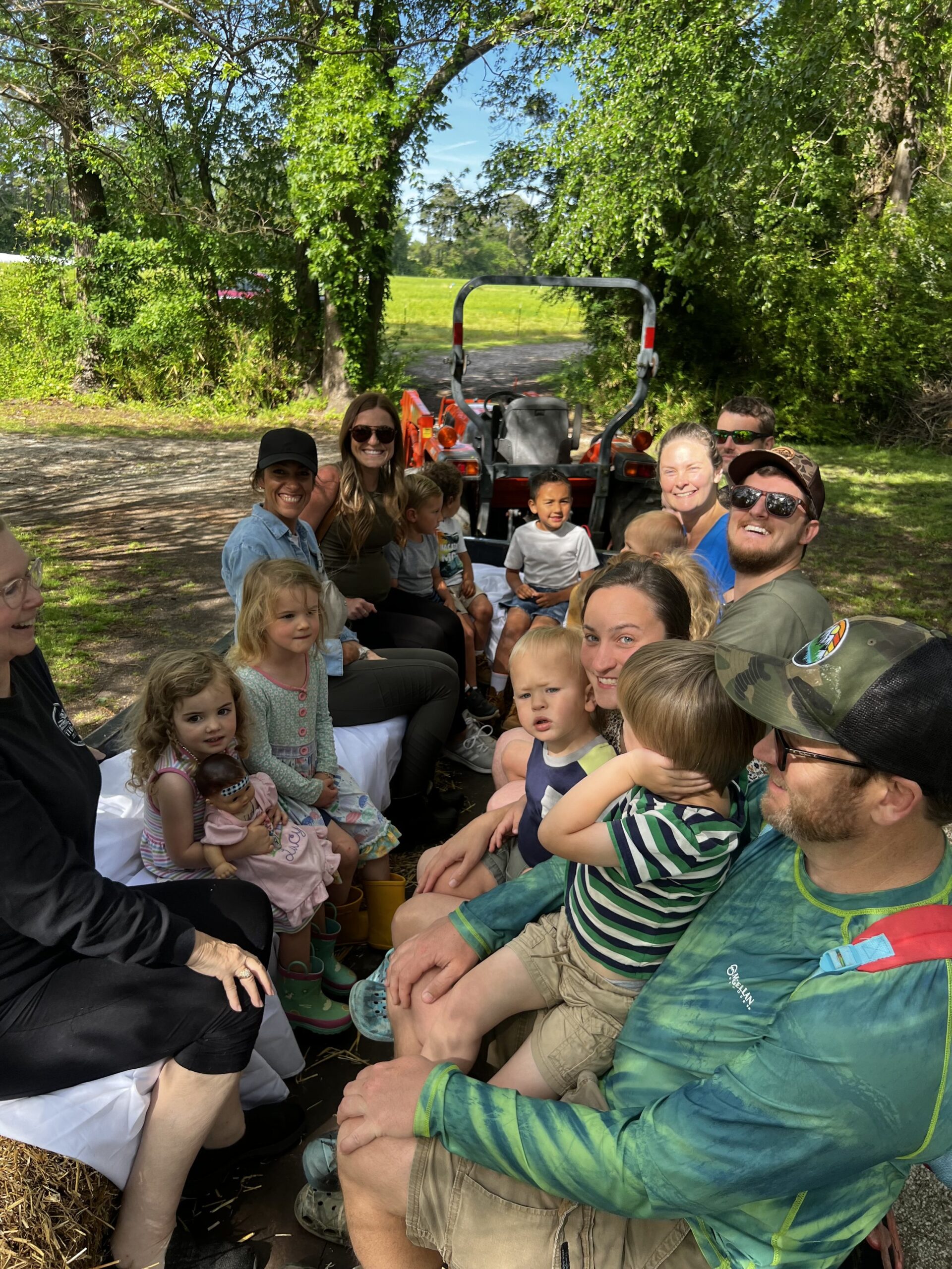 A group of adults and children enjoying a fun outdoor activity while sitting on a tractor ride, surrounded by lush greenery and a sunny atmosphere.
