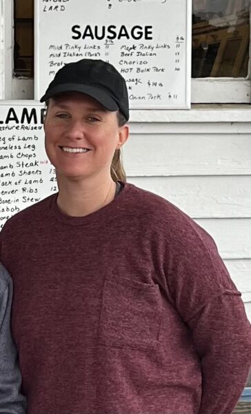 Smiling woman wearing a dark cap and a maroon sweatshirt stands in front of a menu board featuring various meat options, including sausages and lamb dishes, at a food stall or market.