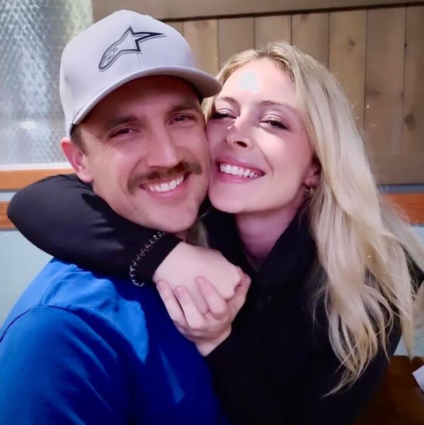 Smiling couple posing together in a cozy indoor setting, with the man wearing a gray cap and a blue shirt, and the woman in a black top, both showing affection and happiness.