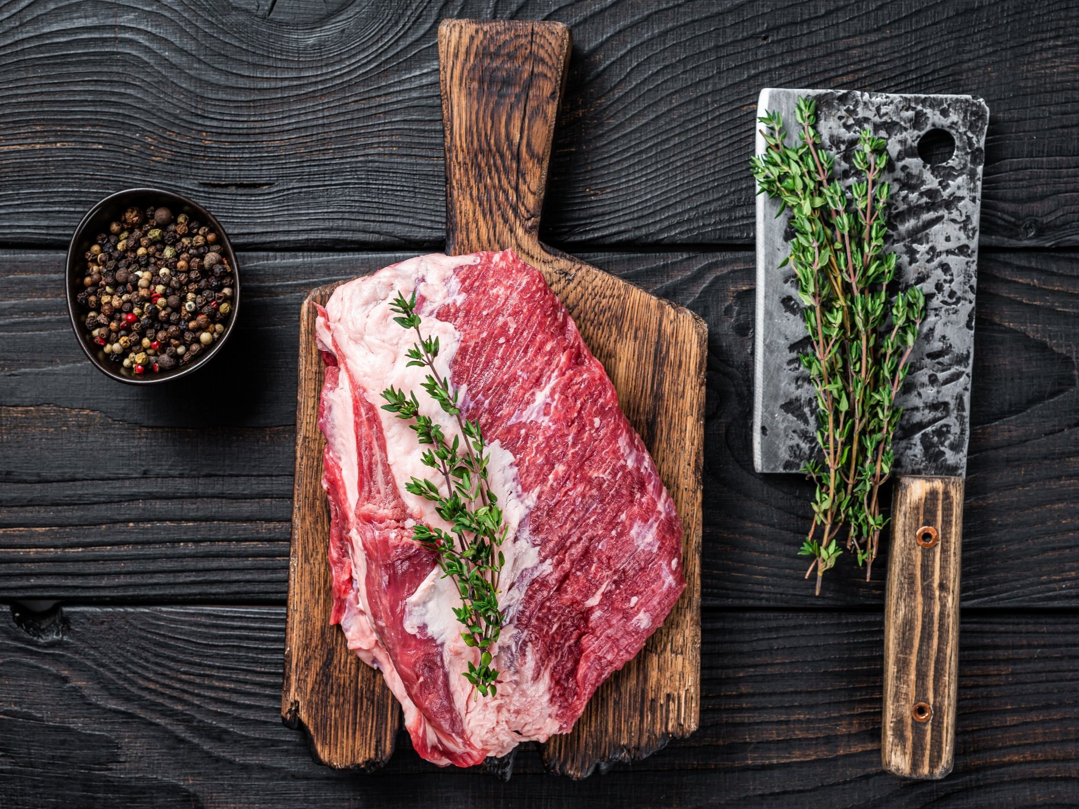 Fresh cut beef brisket on a wooden cutting board, garnished with fresh thyme, accompanied by a bowl of mixed peppercorns and a cleaver on a dark wooden surface.