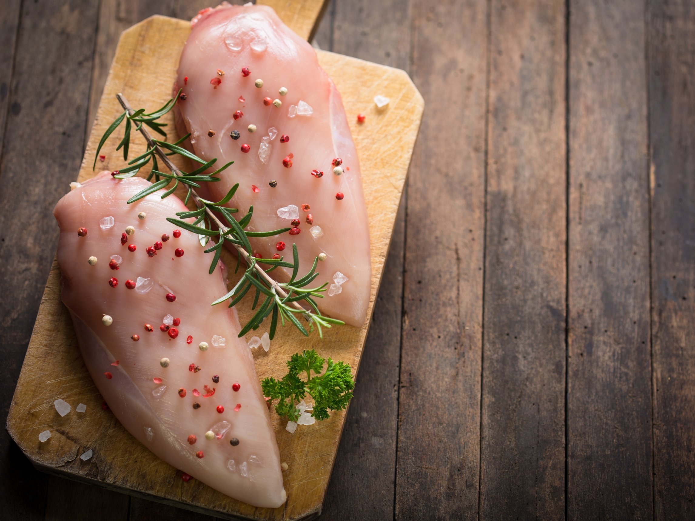 Raw chicken breasts seasoned with colorful peppercorns and fresh rosemary on a wooden cutting board, showcasing a rustic kitchen setting.
