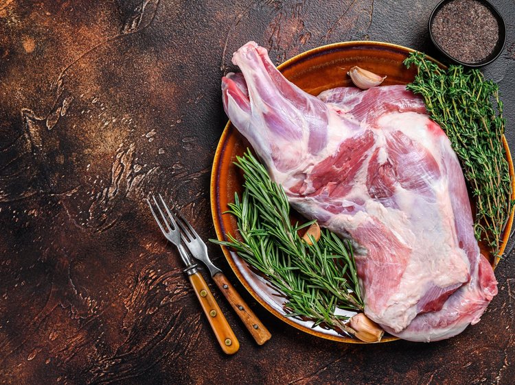 Raw lamb leg displayed on a rustic plate with fresh rosemary, thyme, and garlic, accompanied by two metal forks and a small bowl of black pepper on a textured dark background.