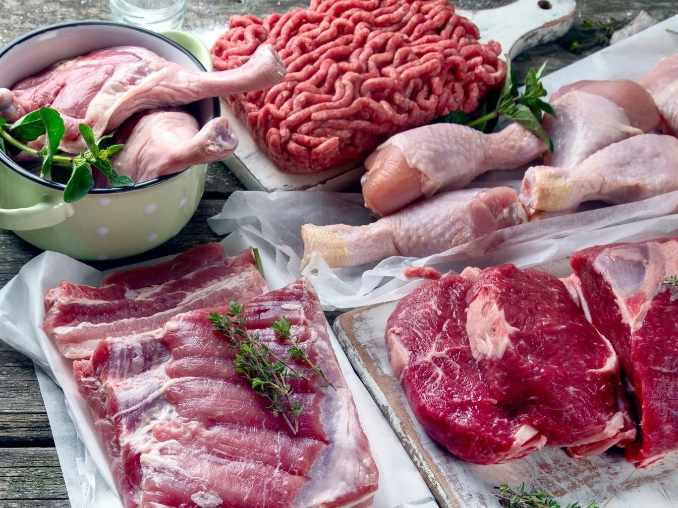 Assorted raw meats displayed on a wooden surface, including chicken legs, ground beef, pork ribs, and steak, garnished with fresh herbs like thyme and mint, ideal for meal preparation and cooking inspiration.