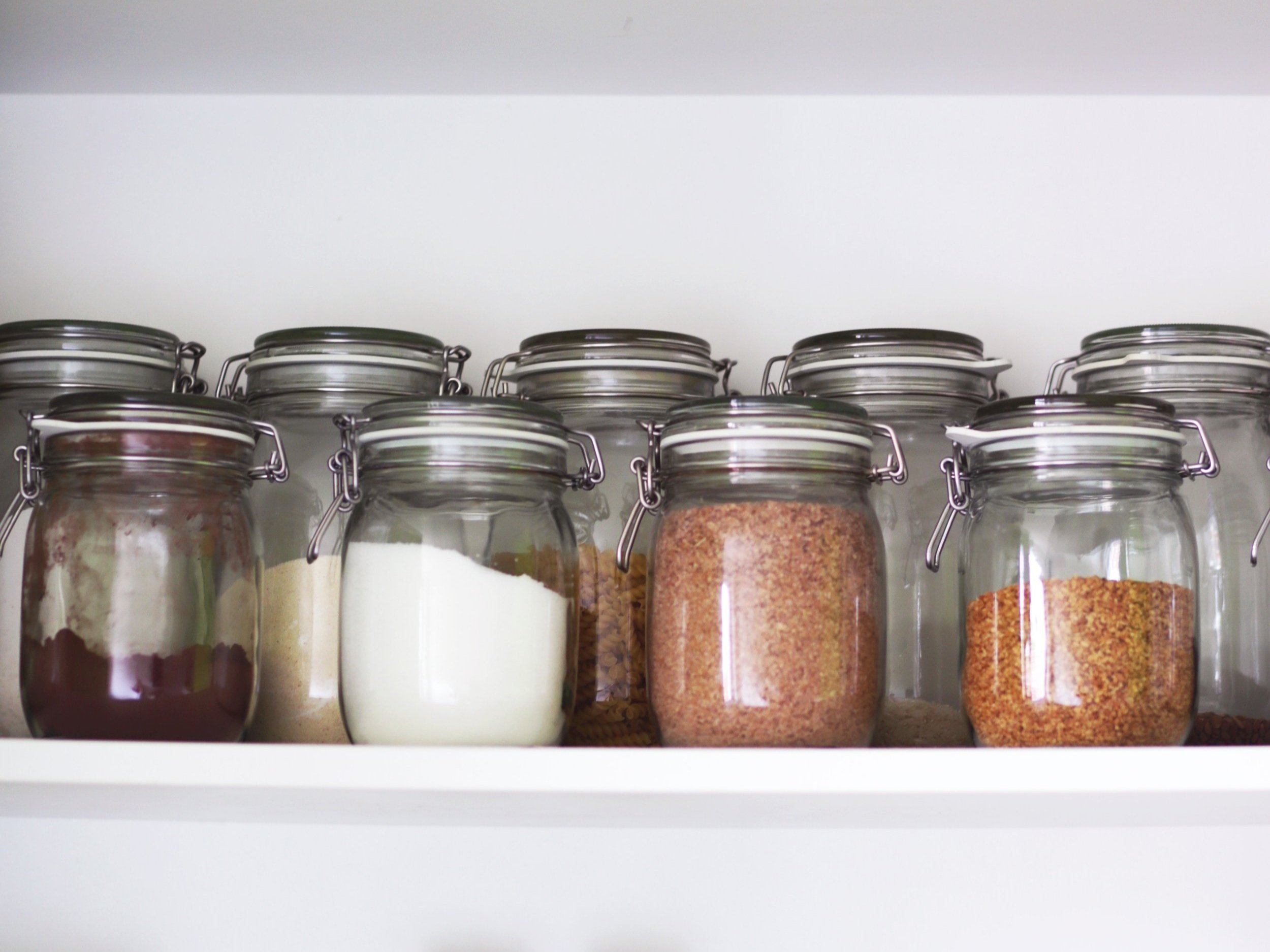 Glass jars filled with various dry ingredients, including sugar, flour, and spices, neatly organized on a white kitchen shelf.