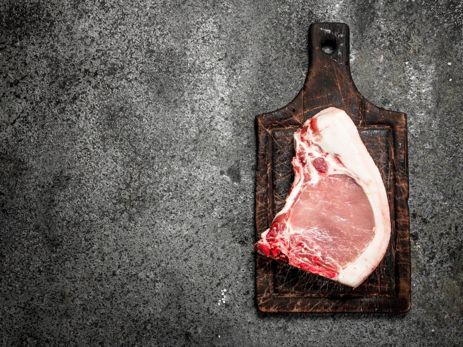 Raw pork chop on a dark wooden cutting board against a textured gray background, highlighting the marbling and freshness of the meat.