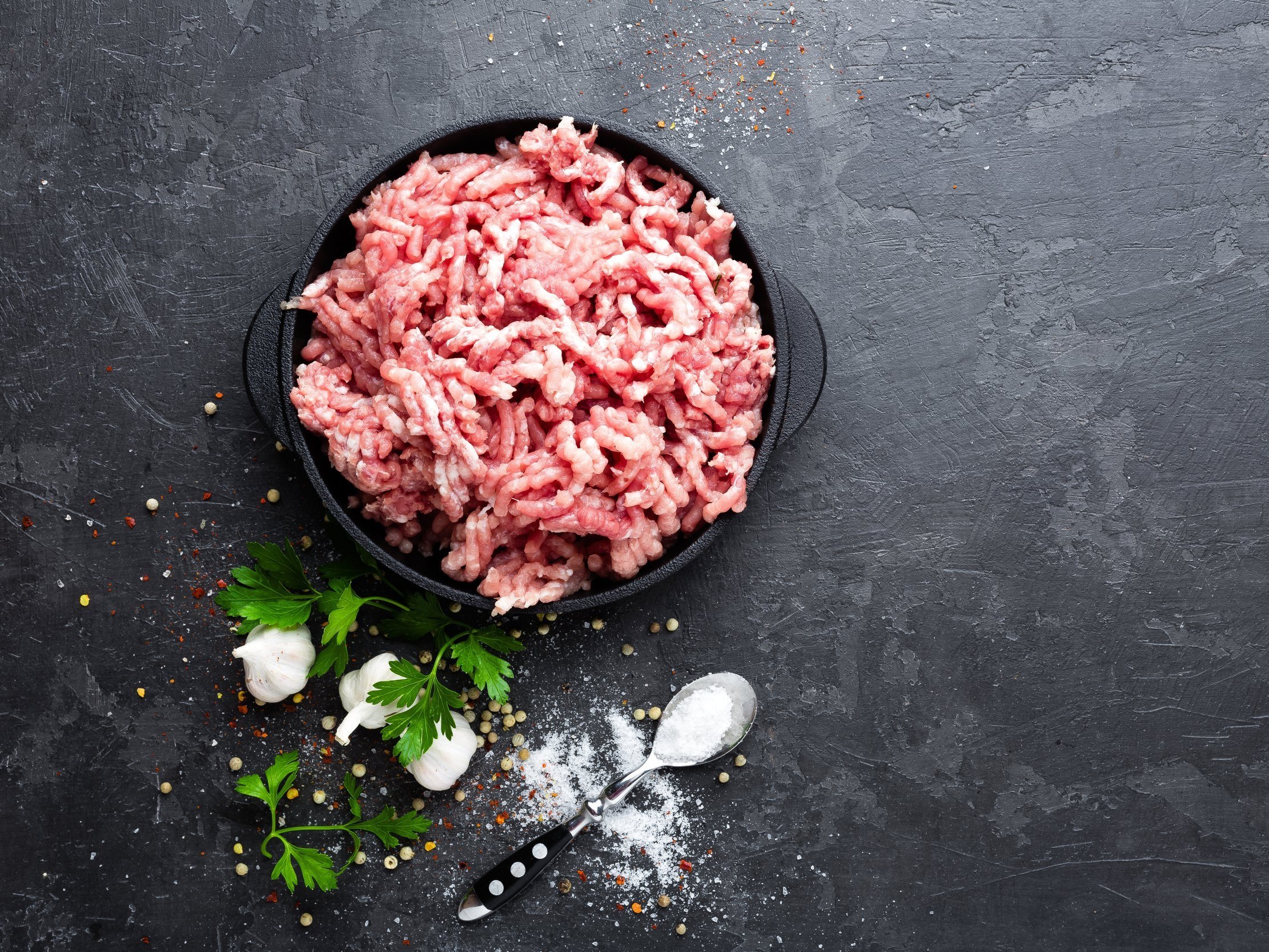 Raw ground meat in a black cast iron skillet, surrounded by fresh parsley, garlic cloves, and scattered spices on a dark textured surface.