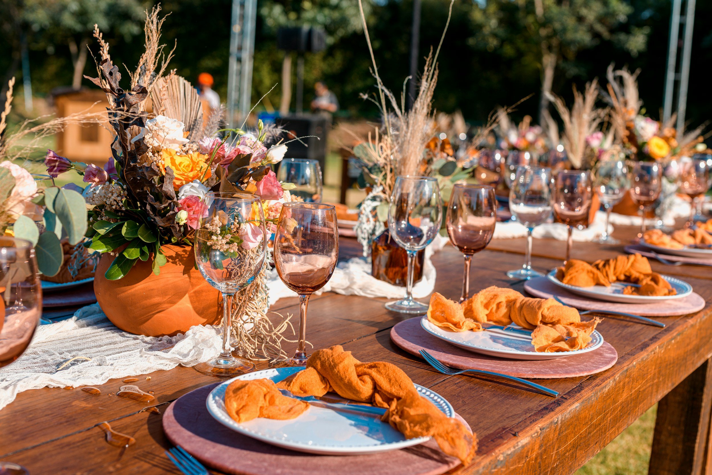 Elegant outdoor dining setup featuring a wooden table adorned with vibrant floral arrangements in terracotta pots, crystal wine glasses, and neatly arranged plates with orange napkins. The scene captures a warm, inviting atmosphere perfect for gatherings and celebrations.